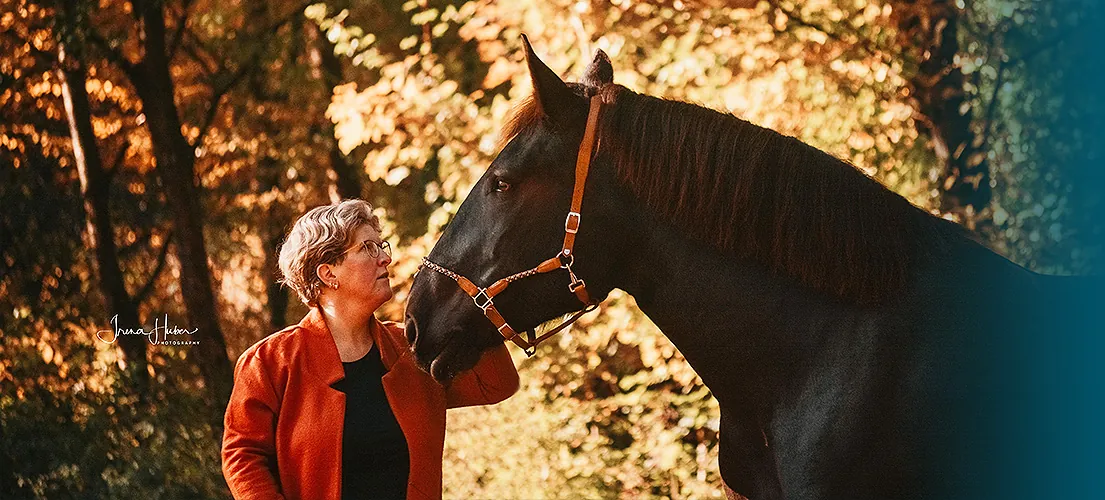 Michaela Sprogis steht im herbstlichen Wald Nase an Nase mit ihrem dunklen Warmblut